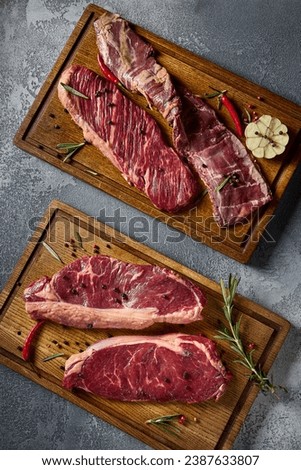 Close-up vertical top-down view, accentuating the intricate marbling of the beef cuts. The raw steaks are artfully arranged on wooden boards, set against a gray canvas.