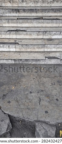 A close-up vertical view of weathered stone stairs with cracks and signs of decay leading to a rough concrete ground surface.