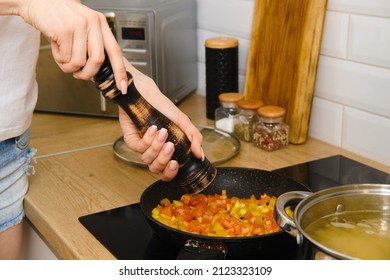 Closeup view of adding black pepper into vegetables in a frying pan. Woman uses pepper mill in the kitchen.