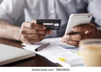 Close-up View Of African Man's Hands Holding Plastic Credit Card And Smart Phone. Dark-skinned Businessman Using Mobile Banking Application On His Gadget While Paying Bills At Cafe Online Via Internet