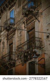 A close-up view of an aged building facade in Catania, Sicily, Italy, featuring multiple ornate iron balconies and wooden window shutters. The architecture displays weathered textures and classical details like corbels and moldings. Electrical wires and a classic street light add to the urban charm. The natural light highlights the building's historical character, creating a rich, melancholic atmosphere. Ideal for travel publications, historical documentaries, and architectural studies exploring European heritage.