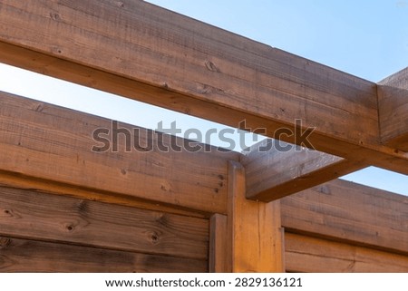Close-up view of aged, dark brown wooden beams forming an outdoor pergola structure with a bright sky visible through the gaps