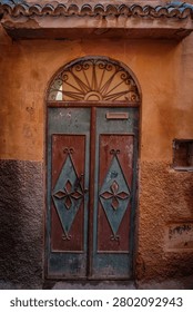 A close-up view of an aged, distressed double door in a traditional Moroccan style, featuring peeling teal and burgundy paint with diamond and quatrefoil-like patterns. Above the door, an arched wrought iron transom window with scrollwork is set into an ochre-colored plaster wall with decorative blue-green roof tiles.