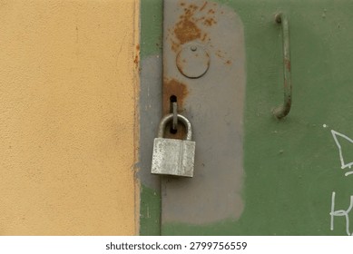 Close-up view of an aged silver padlock hanging from a hasp on a green metal door next to a beige painted wall. The door shows patches of rust, chipped paint