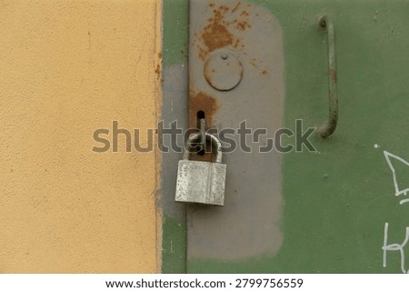 Close-up view of an aged silver padlock hanging from a hasp on a green metal door next to a beige painted wall. The door shows patches of rust, chipped paint