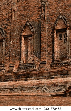 Close-up view of an ancient red brick wall featuring pointed arch windows at the historic ruins of Wat Kudi Dao in Ayutthaya, Thailand. The weathered facade shows old stucco details in bright daylight