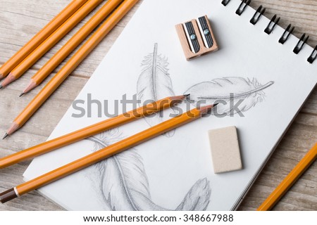 Close-up view of artist's or designer's table. Pencils, sharpner and eraser laying on sketch book with hand-drawn feathers