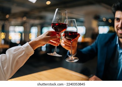 Close-up view from back of unrecognizable young woman clinking glasses of red wine with handsome smiling man sitting at table in restaurant at evening. Happy young couple enjoying nice romantic dinner