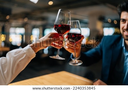 Close-up view from back of unrecognizable young woman clinking glasses of red wine with handsome smiling man sitting at table in restaurant at evening. Happy young couple enjoying nice romantic dinner