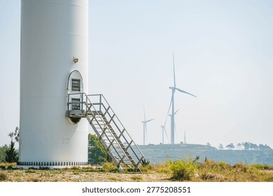 A close-up view of the base of a white wind turbine featuring a metal staircase leading to an oval door, with more turbines visible on a distant hazy ridge.