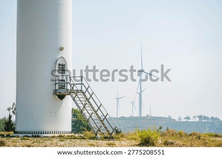A close-up view of the base of a white wind turbine featuring a metal staircase leading to an oval door, with more turbines visible on a distant hazy ridge.