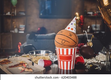 Close-up View Of Basketball Ball With Funny Face In Popcorn Box And Messy Table After Party 