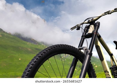 Close-up view from below of the handlebars and part of the wheel of a mountain bike standing against the backdrop of green hills and cumulus clouds on a sunny day. Vehicle