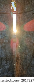 Close-up view between two large tree trunks with rough textured bark, revealing a narrow gap where sunlight shines brightly. The glowing rays create a lens flare effect with reddish and yellowish hues, suggesting sunrise or sunset. The contrast between the rugged bark and radiant light highlights the natural beauty of illumination through trees, perfect for themes of nature, tranquility, spirituality, and outdoor landscapes.