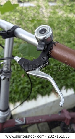 Close-up view of a bicycle handlebar featuring a silver bell, metal brake lever, brown grip, and black mechanical components. The bike is photographed outdoors with green vegetation in the background. Suitable for cycling, transportation, bicycle accessories, mechanical parts, maintenance, commuting, and outdoor activity concepts.
