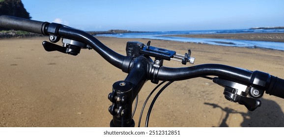 A close-up view of a bicycle handlebar with grips, a bell, and a phone holder. The bicycle is parked on a sandy beach with the ocean in the background.

 - Powered by Shutterstock