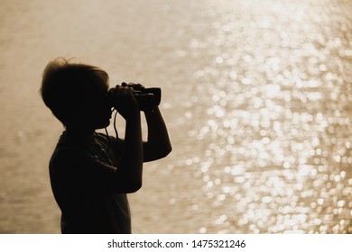 Closeup view of black silhouette of little kid standing outdoor at blurry golden river water background. Boy looking in old vintage binocular. Horizontal color photography.