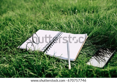 Close-up view of blank notebook with pencil, eyeglasses and smartphone on green grass