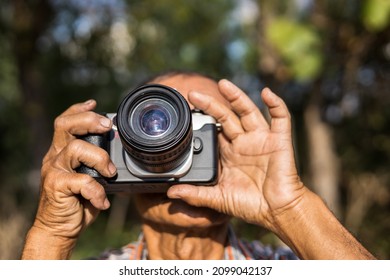 Close-up view of both hands of an elderly Thai man holding a film camera, focusing his lens to photograph something during the day in a forest garden in a rural area of Thailand.