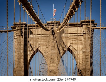 A close-up view of the Brooklyn Bridge, showcasing its iconic stone towers and suspension cables against a clear blue sky. An American flag is visible at the top of one tower. - Powered by Shutterstock