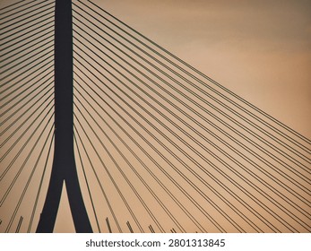 A close-up view of the cables and pylon of a cable-stayed bridge against a sunset sky. The symmetrical geometric patterns of the suspension cables create a stark silhouette at dusk.