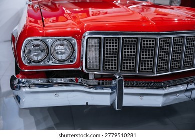 Close-up view captures front end of a vibrant red vintage car. Chrome grille and dual headlights shine under bright lighting, reflecting its well-maintained condition in a showroom-like setting.