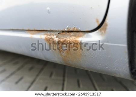 Close-up view of a car lower side panel with severe rust and corrosion, showing flaking paint and oxidized metal, captured outdoors on a paved surface.