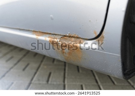 Close-up view of a car lower side panel with severe rust and corrosion, showing flaking paint and oxidized metal, captured outdoors on a paved surface.