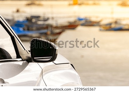 A closeup view of a car side mirror parked near a coastal harbor, with fishing boats softly blurred in the background during golden hour