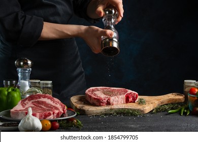 Close-up view of chef pours black pepper on raw steak on wooden chopped board. Backstage of preparing grilled pork meat at restaurant kitchen on dark blue background. Frozen motion. - Powered by Shutterstock