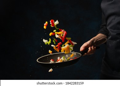 Close-up view of chef's hand throws up frying mix of colored vegetables above the pan on dark blue background. Backstage of cooking meal. Frozen motion. Food banner concept. - Shutterstock ID 1842283159