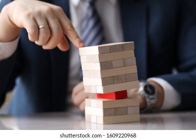 Close-up view of company leader pressing on wooden tower on one hand. Skewed pyramid of light blocks with one red brick in the middle. Risk and vulnerable situation concept