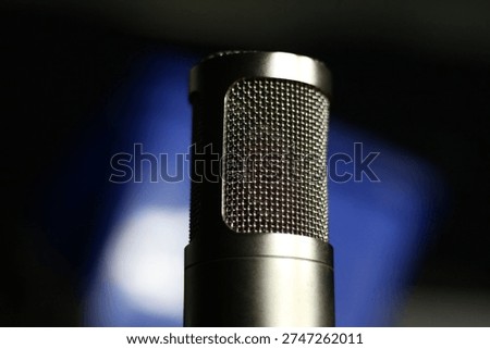 Close-up view of a condenser microphone with a metallic mesh grille, placed in a studio setting with a softly blurred blue background. The image highlights the technical details and clean construction of professional audio equipment used for recording, podcasting, broadcasting, voice work, music production, and sound engineering. Suitable for commercial themes related to media creation, studio workflow, and modern audio technology.