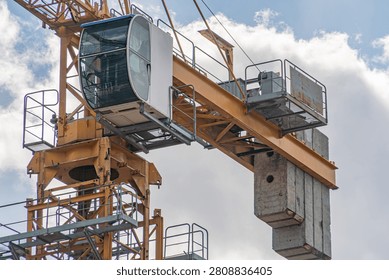 Close-up view of construction crane, featuring operator's cab with glass enclosure, robust metal framework, boom section, concrete pillar attachment, partly cloudy sky background, natural sunlight