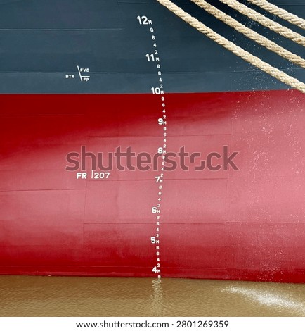 Close-up view of a container ship hull showing draft marks and mooring lines alongside the vessel. Maritime transportation details captured from the ship’s side with industrial nautical elements visible
