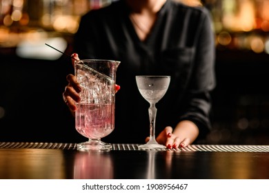 close-up view of crystal glass and mixing cup with pink drink on bar counter. Female bartender in the background