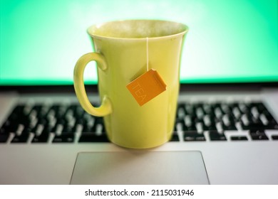 Closeup View Of A Cup Of Hot Beverage And A Laptop With Green Screen In Background. Steaming Yellow Mug On Computer Keyboard. Remote Workstation. Smart Working, Home Working And Homeschooling Concept