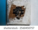 A close-up view of a damaged mechanical or electrical component embedded in a weathered wall. The corroded metallic parts and deteriorated surroundings emphasize decay and industrial aging