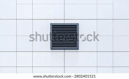 A close-up view of a dark gray square ventilation grille set against a clean white tiled wall, highlighting a common architectural detail.