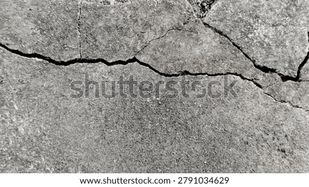 Close-up view of deep, irregular cracks spreading across a rough, textured gray concrete surface. The fractures form a jagged, branching pattern, highlighting signs of aging, structural damage, wear, and weathering on solid cement material. Monochrome gray tones emphasize the raw, gritty texture and broken, uneven lines of the cracked pavement or wall.