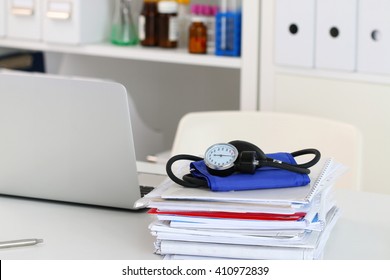 Closeup View Of Doctor Working Table. Laptop, Manometer And Heap Of Documents Lying On Desktop, Physician Office. Healthcare And Medical Concept.