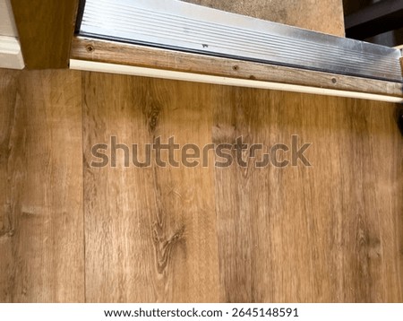 Close-up view of a door threshold where a light brown wood floor meets a raised metal edge and a concrete step. The worn wooden transition strip and screw-mounted metal trim show subtle texture and