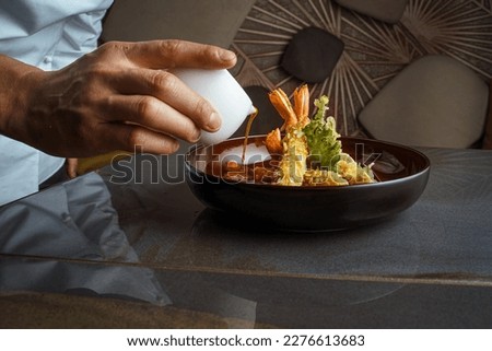 Close-up view of an elegant black bowl filled with tempura shrimp and vegetables, topped with sweet and sour sauce poured by the chef's hand