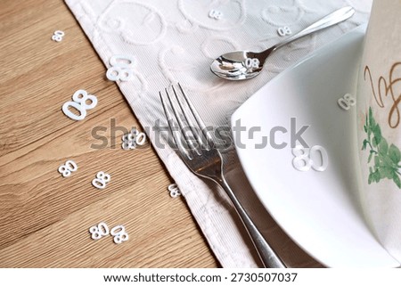 Close-up view of an elegant table setting decorated with white number eighty confetti scattered on a textured table runner and wooden table surface. A plate, fork, spoon, and folded napkin create a refined dining atmosphere. The image represents an 80th birthday, anniversary, or milestone celebration in a restaurant or private event setting, with ample copy space for design and editorial use.