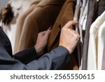 Close-up view of elegant woman in black jacket holding brown coat and choosing clothes from hangers in clothing store. Soft focus. Copy space. Fashion and retail business theme.