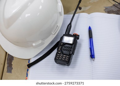 Close-up view of essential construction site equipment: a white hard hat, a two-way radio, and an open notebook with a pen, suggesting planning and communication.