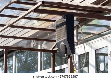 Close-up view of exterior intercom with mesh grille and circular speaker mounted beneath a glass-and-metal canopy near building doors, reflecting trees and sky and forming a clean contemporary archite