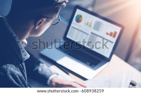 Closeup view of finance market analyst in eyeglasses working at sunny office on laptop while sitting at wooden table.Businessman analyze stock report on notebook screen.Blurred background,horizontal