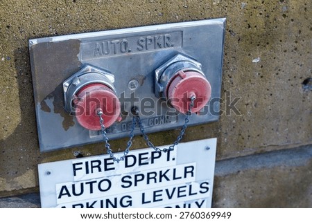 Close-up view of a fire department automatic sprinkler siamese connection panel mounted on a weathered concrete exterior wall. The stainless steel panel is stamped with Auto. Spkr. and features two red-capped fire department connection inlets connected by a metal chain to prevent cap loss. The chrome couplings show signs of age and patina on the metal plate. Below the connection panel, a white sign reads Fire Dept. Auto Sprklr Parking Levels Only, indicating the sprinkler system serves the parking structure. The concrete wall surface shows weathering, mineral staining, and aging. This type of fire suppression infrastructure is a standard requirement in commercial and institutional buildings, providing external access points for firefighters to supplement the building's automatic sprinkler system with additional water pressure during emergencies