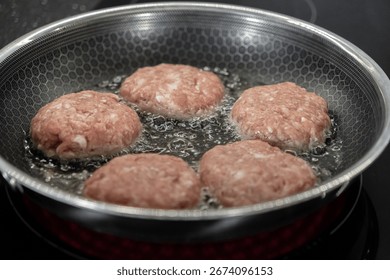 Close-up view of five raw cutlets bubbling in hot oil inside a textured non-stick frying pan, showcasing the initial stage of home meal preparation